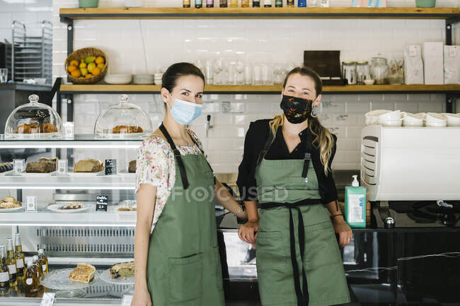 Partners wearing face mask and apron standing by counter at coffee shop — Stock Photo