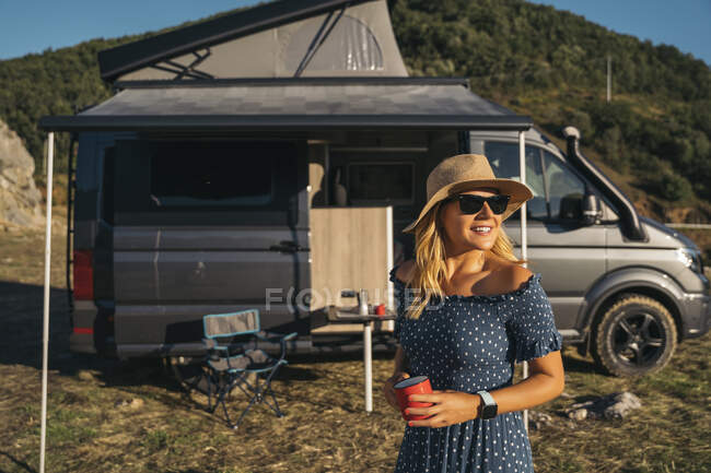 Femme souriante avec tasse de café regardant loin contre le camping-car par une journée ensoleillée — Photo de stock