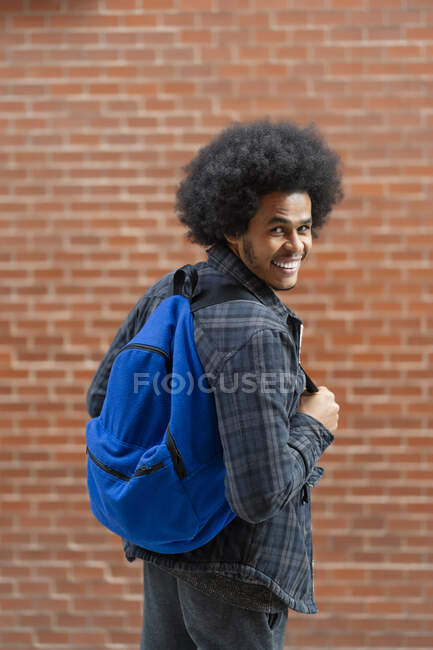 Веселий молодий чоловік з блакитним рюкзаком стоїть перед цегляною стіною. — стокове фото