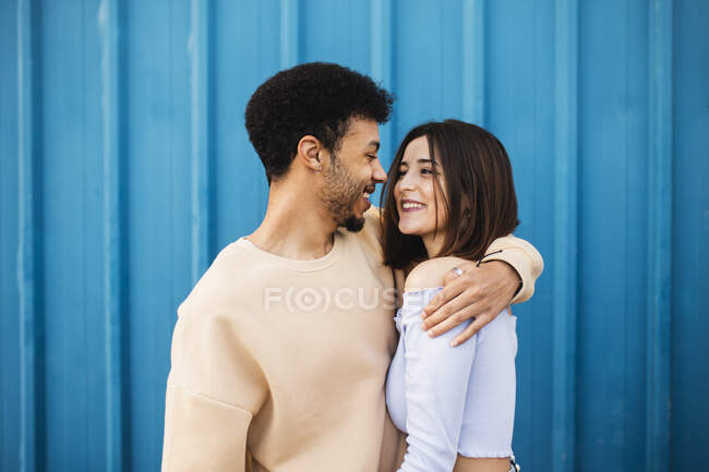 Sonriente joven abrazando a la mujer mientras está de pie contra la pared azul - foto de stock