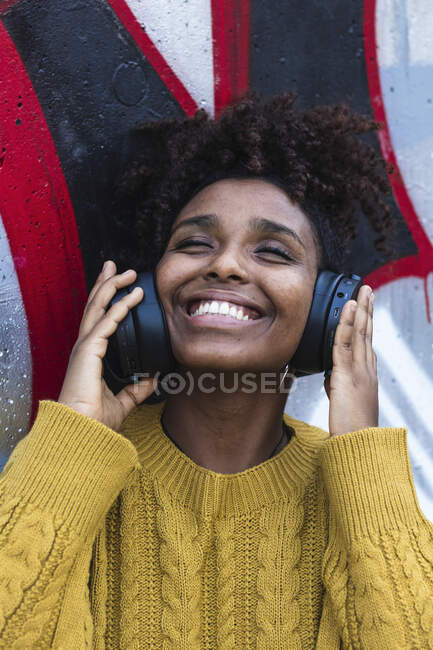 Joyeux jeune femme avec les yeux fermés écouter de la musique à travers des écouteurs contre le mur peint — Photo de stock