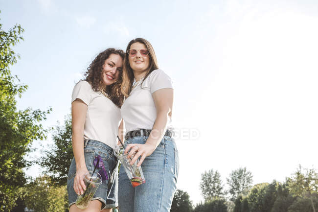 Lächelnde schöne Frauen stehen an sonnigen Tagen mit Einwegbechern im Park gegen den Himmel — Stockfoto