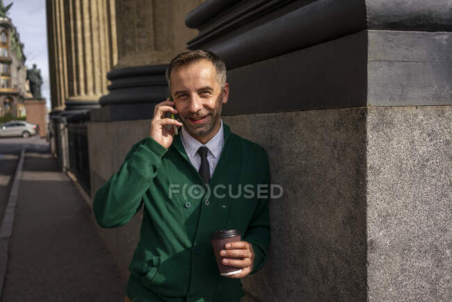 Улыбающийся бизнесмен разговаривает по мобильному телефону, опираясь на колонну Казанского собора в Санкт-Петербурге, Россия — стоковое фото