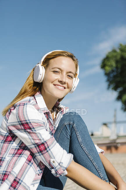 Donna sorridente che ascolta musica sulla cuffia seduta sul sentiero durante la giornata di sole — Foto stock