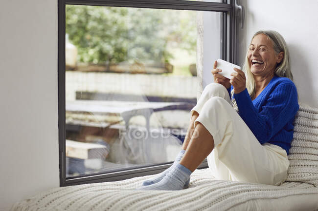 Смеющаяся взрослая женщина держит мобильный телефон, сидя у окна дома — стоковое фото
