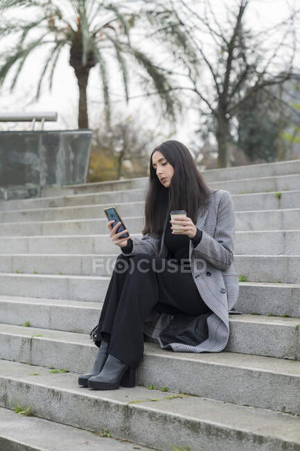Mujer de negocios de moda usando el teléfono móvil mientras toma café en la escalera - foto de stock