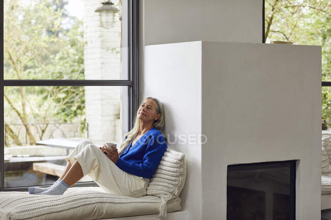 Mature woman with coffee cup day dreaming while leaning on wall of fireplace at home — Stock Photo
