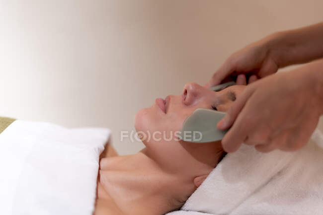 Esteticista sosteniendo gua sha en las mejillas del cliente femenino en el spa de salud - foto de stock