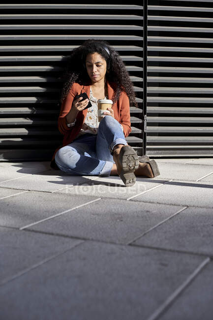 Frau mit Smartphone hält Kaffeetasse während sie gegen Verschluss sitzt — Stockfoto