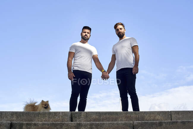 Гей пара тримає руки, стоячи на сходах проти неба — стокове фото