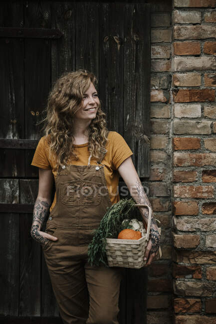 Улыбающаяся женщина держит корзину с овощами у стены — стоковое фото