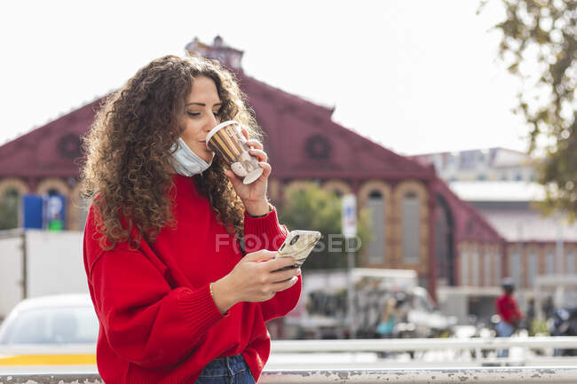 Mujer joven bebiendo café mientras usa teléfono móvil en la ciudad durante COVID-19 - foto de stock