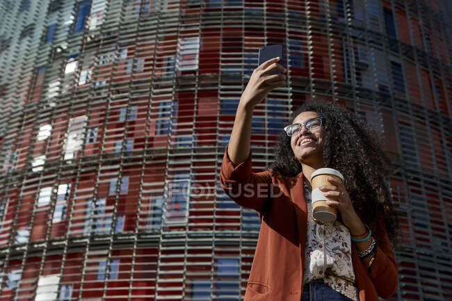 Sorrindo jovem mulher fazendo videochamada no telefone inteligente enquanto está contra a construção durante o dia ensolarado — Fotografia de Stock