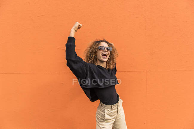 Веселая женщина с поднятыми руками, стоящая у оранжевой стены — стоковое фото