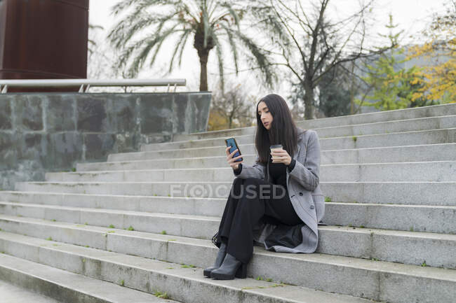 Жінка-підприємець з чашкою кави багаторазового використання мобільного телефону на сходах — стокове фото