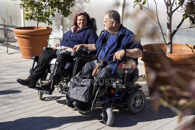 Женщина-инвалид и мужчина держатся за руки, сидя в инвалидном кресле в солнечный день — стоковое фото