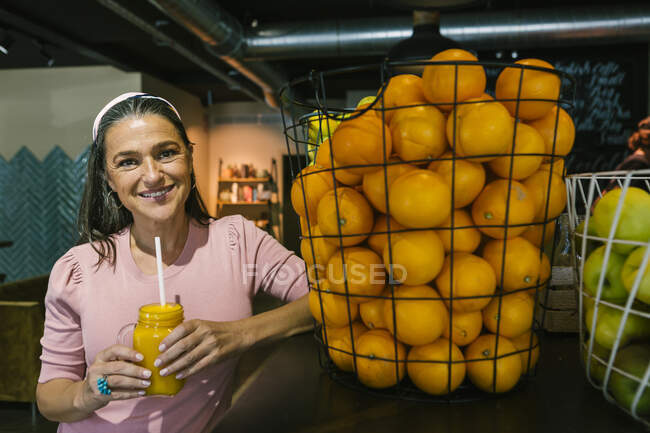 Посмішка дорослої жінки, яка тримає свіжий сік у глечику для каменю, стоячи біля апельсинів у металевому кошику в кафе. — стокове фото