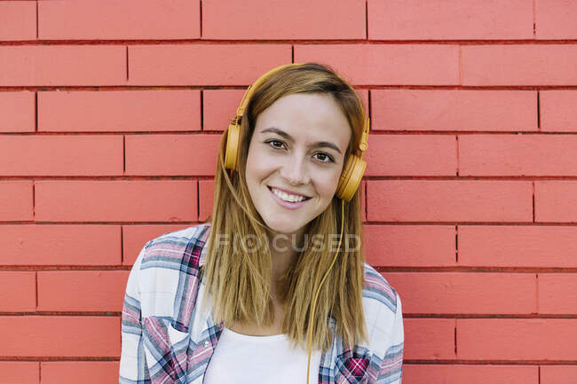 Donna sorridente che ascolta musica attraverso le cuffie contro il muro di mattoni — Foto stock