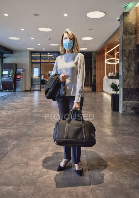 Frau trägt Gesichtsmaske, während Tasche in Hotellobby steht — Stockfoto