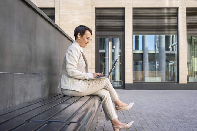 Femme utilisant un ordinateur portable tout en étant assis sur le banc contre le bâtiment — Photo de stock