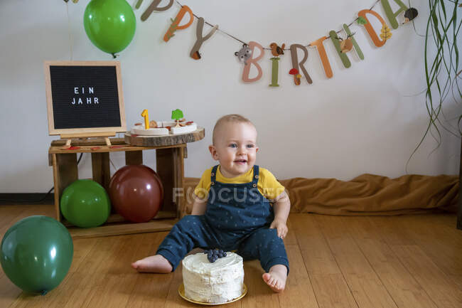 Smiling baby boy sitting with cake on floor against decoration at home — Stock Photo