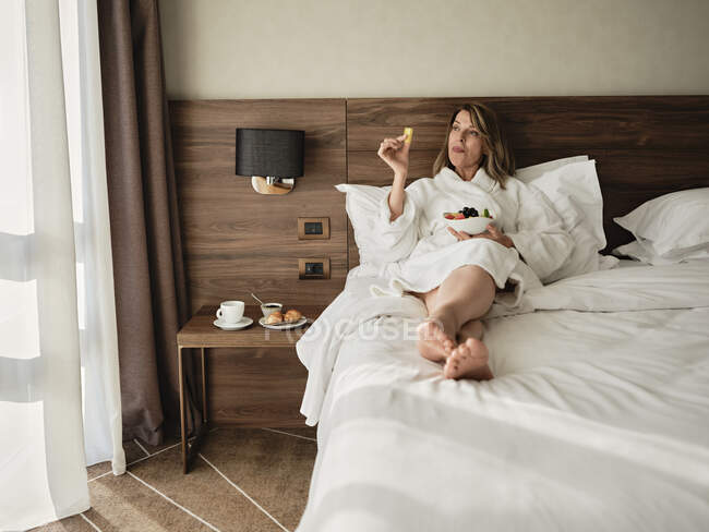 Mulher sênior comendo frutas frescas no café da manhã enquanto relaxa na cama e olha para o quarto de hotel — Fotografia de Stock