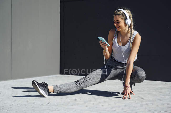 Улыбающаяся спортсменка в наушниках с помощью мобильного телефона, приседающая на тропинке — стоковое фото