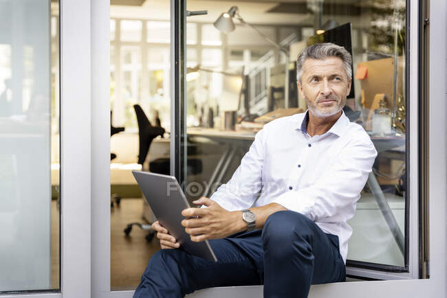 Pensativo hombre de negocios con tableta digital mirando hacia otro lado mientras se apoya en la puerta de la oficina - foto de stock