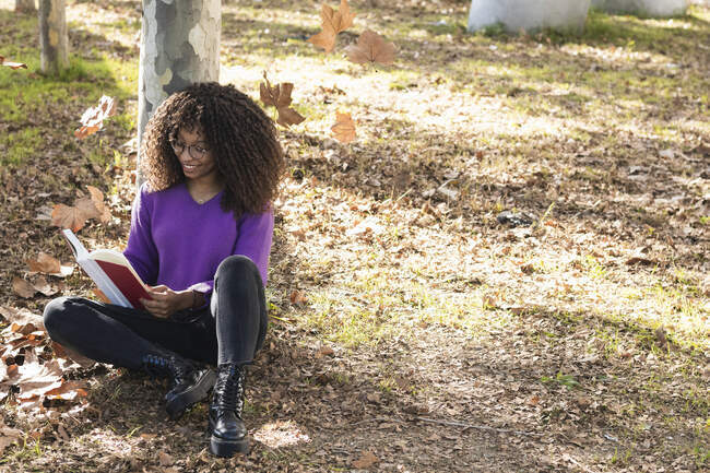 Усмішка модна жінка читає книжку, спираючись на стовбур дерева в громадському парку. — стокове фото