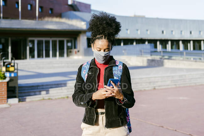 Estudiante usando mascarilla protectora usando teléfono inteligente mientras está parado en la calle de la ciudad - foto de stock