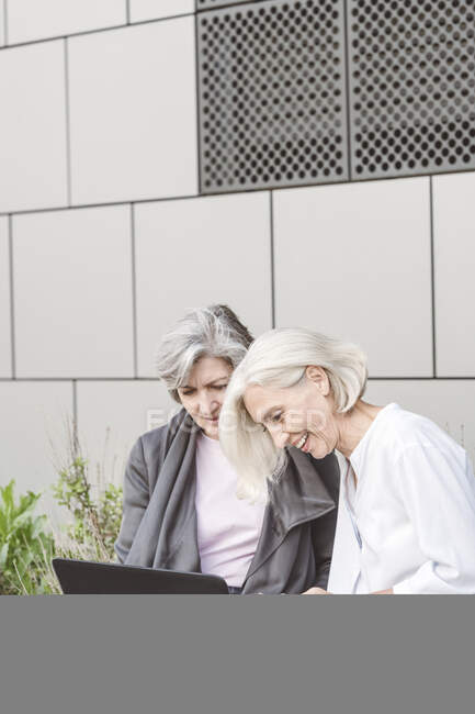 Smiling businesswomen working on laptop while sitting against building — Stock Photo