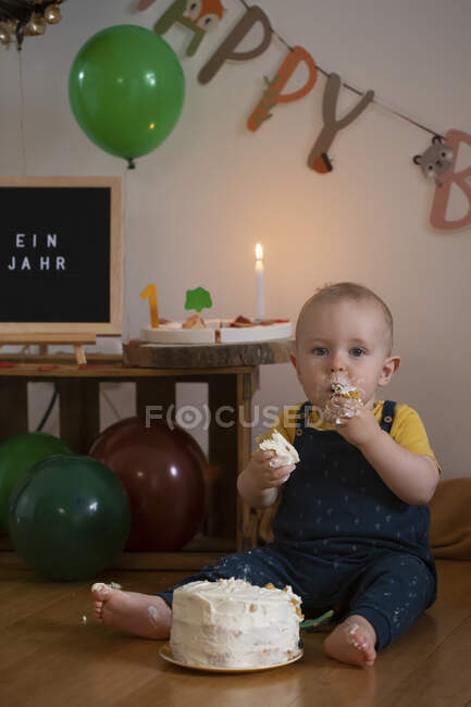 Hungry baby boy eating cake of his first birthday while sitting against birthday decoration at home — Stock Photo