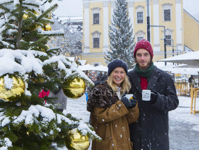 Young couple holding cups with mulled wine while standing by Christmas tree in city — Stock Photo