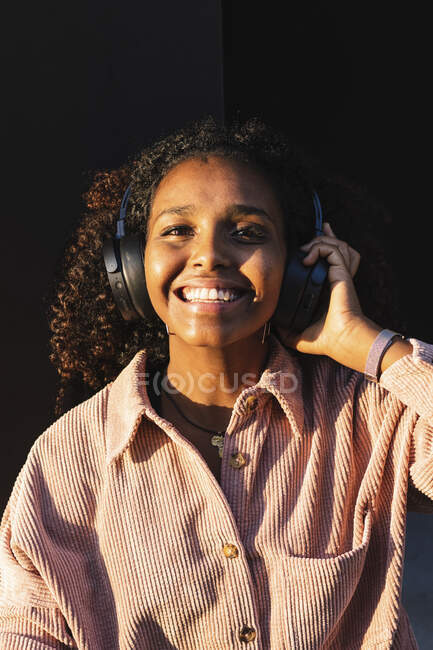 Щаслива модна молода жінка слухає музику проти чорної стіни в сонячний день — стокове фото