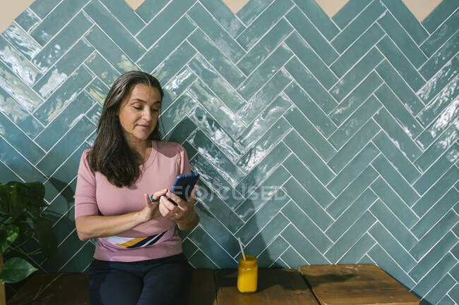 Жінка використовує смартфон, сидячи з соком проти бірюзової стіни в барі — стокове фото