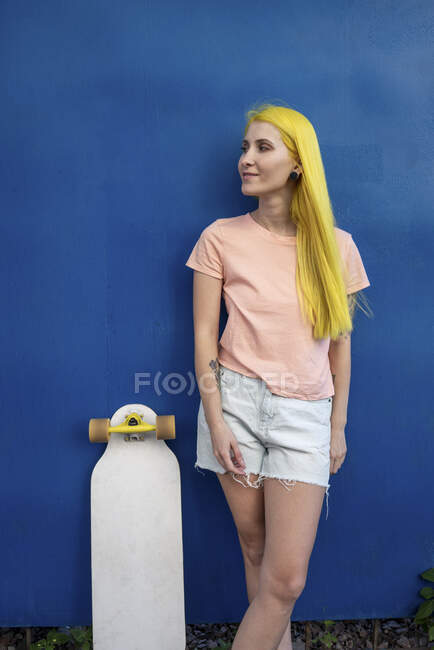 Jeune femme avec planche à roulettes debout contre le mur bleu — Photo de stock