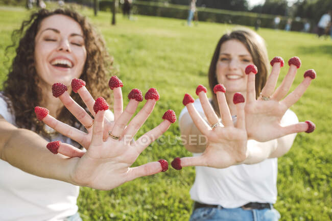 Juguetonas amigas disfrutando con frambuesas frescas en los dedos en el parque durante el día soleado - foto de stock