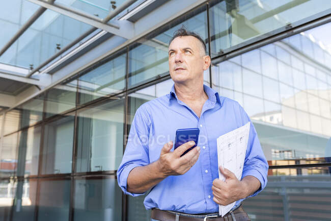 Thoughtful businessman holding smart phone and document while looking away in city during pandemic — Stock Photo