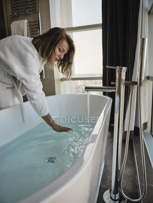 Mulher sênior se curvando enquanto examina a temperatura na banheira no hotel de luxo — Fotografia de Stock