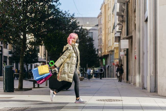 Mulher usando máscara facial correndo com sacos de compras no caminho da cidade — Fotografia de Stock