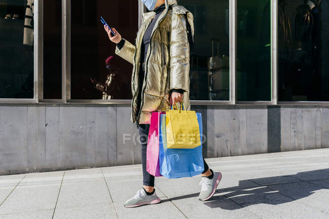 Молода жінка з захисною маскою для обличчя несе сумки з магазином, користуючись мобільним телефоном. — стокове фото