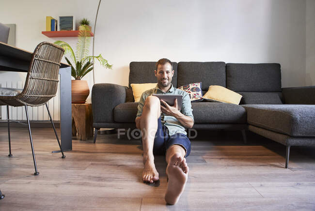 Homem sorridente sentado no chão com tablet digital contra sofá em casa — Fotografia de Stock