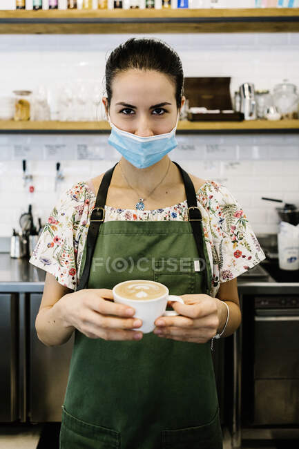 Молода жінка в масці і фартух з чашкою кави стоїть у кав'ярні. — стокове фото