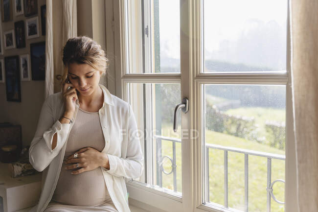 Young pregnant woman with hand on stomach talking through smart phone at home — Stock Photo