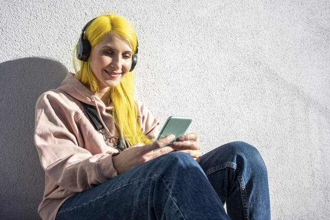 Junge Frau mit Smartphone beim Musikhören über Kopfhörer sitzt an sonnigem Tag gegen Wand — Stockfoto
