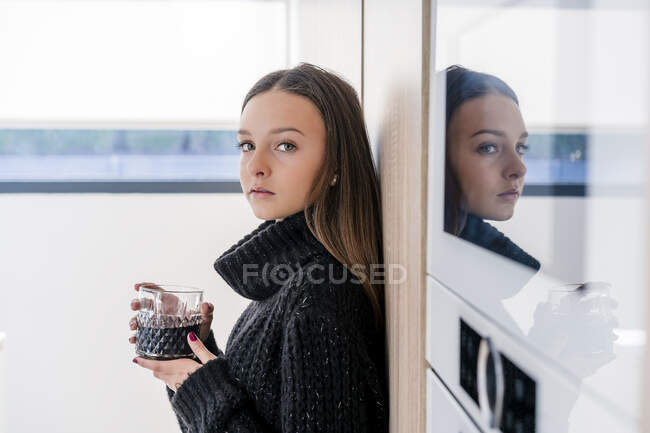 Serious teenage girl with drink standing by appliance in kitchen — Stock Photo