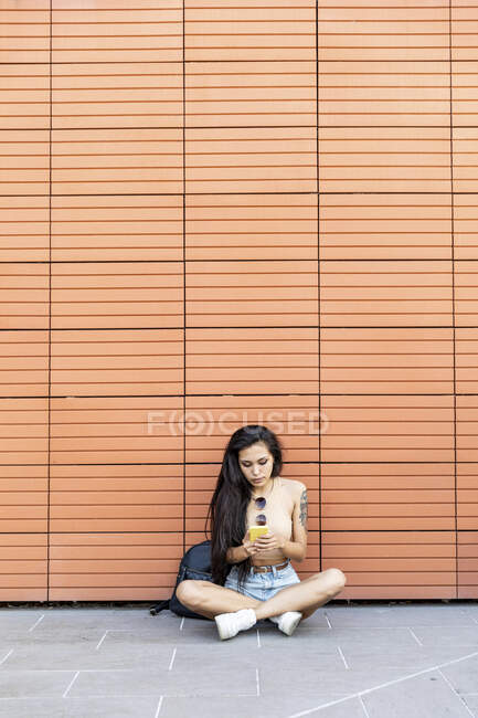 Young woman using mobile phone while sitting against wall — Stock Photo