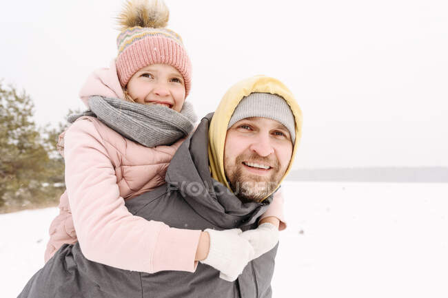 Sonriente padre piggybacking hija en nieve tierra contra claro cielo - foto de stock