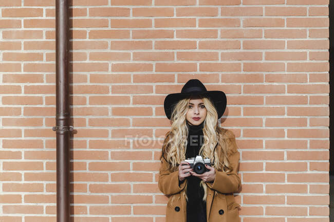 Світла жінка тримає фотоапарат, стоячи проти цегляної стіни в сонячний день. — стокове фото