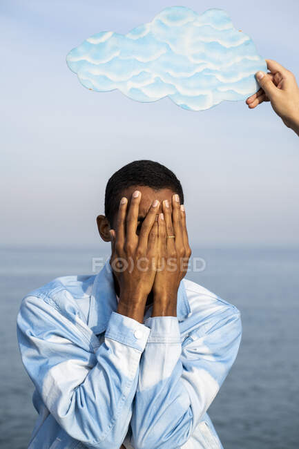 Man covering face while standing under male hand holding cloud cut out against sky - foto de stock